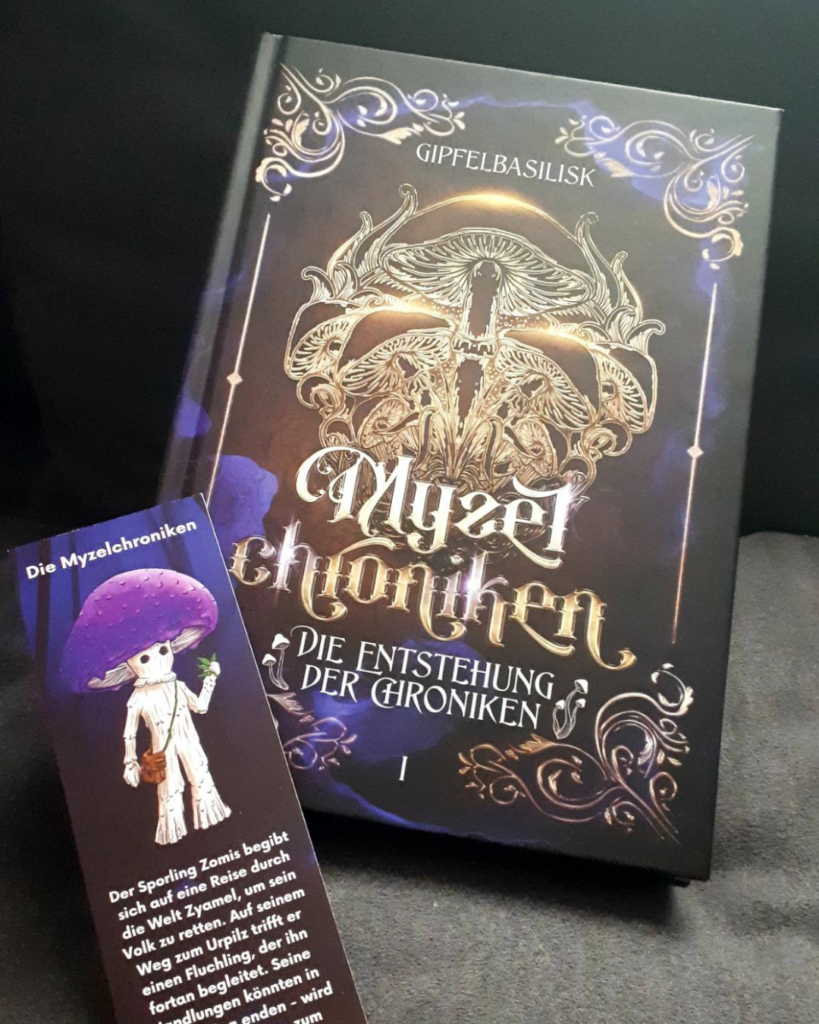 Buch "Myzelchroniken - Die Entstehung der Chroniken" steht auf grauem, weichem Untergrund, ein Lesezeichen mit der Zeichnung eines humanoiden Pilzwesens ist daran angelehnt.