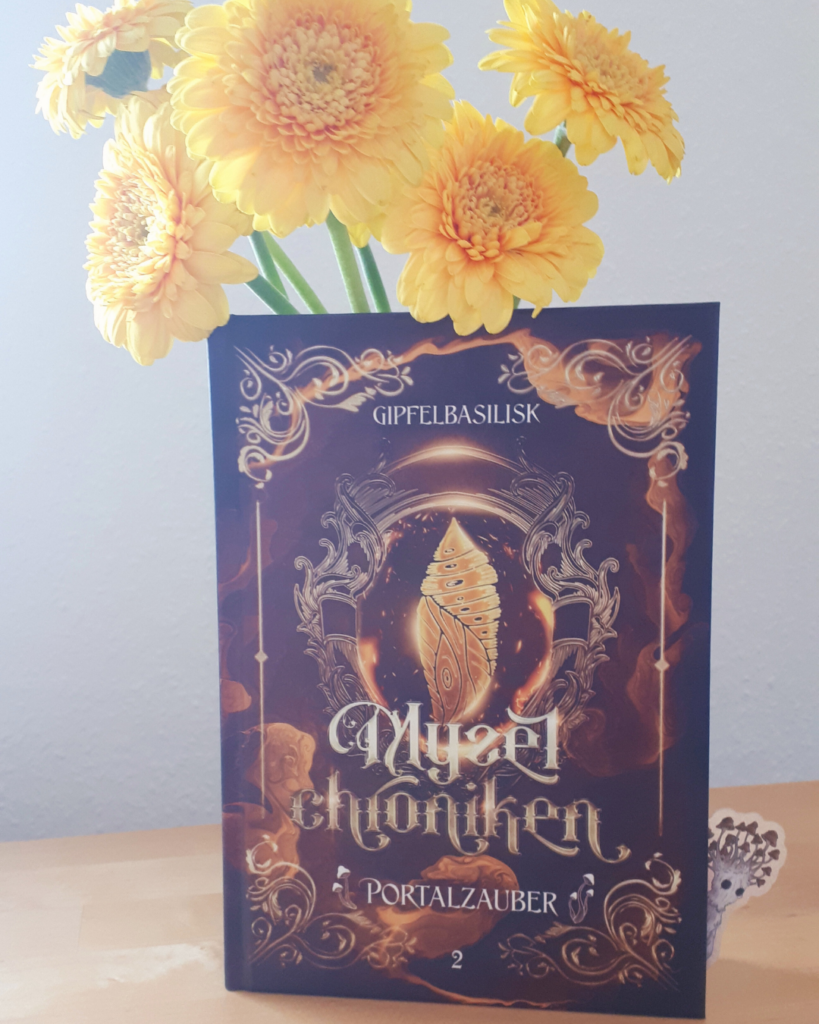 Buch "Myzelchroniken - Portalzauber" steht auf einer Holzoberfläche vor gelb blühenden Blumen, ein glitzernder Sticker eines humanoiden Pilzwesens ragt aus dem Buch