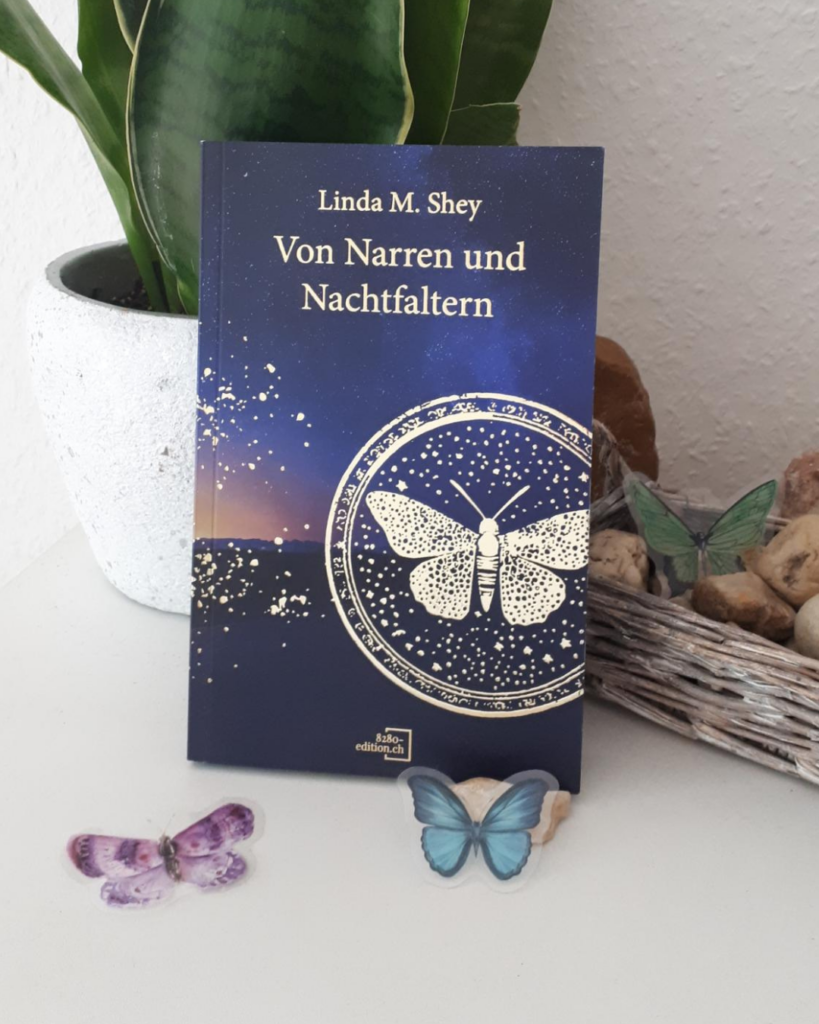Buch "Von Narren und Nachtfaltern" an einen Pflanztopf angelehnt, daneben ein Körbchen mit Steinen, drei Deko-Falter sind um das Buch herum angeordnet